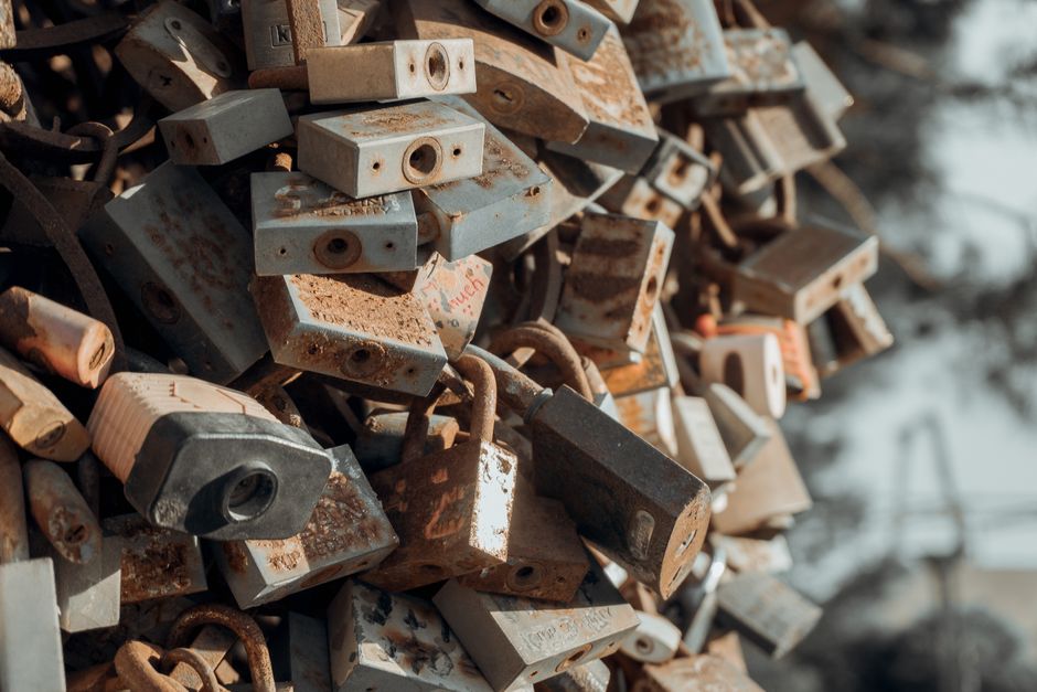 A close-up view of a large pile of rusty metal padlocks, some attached to rusted chains, with various sizes and shapes. The padlocks appear weathered, with visible rust and corrosion on their surfaces, and some have faded or peeling paint. The image is taken outdoors, with a blurred background showing hints of natural surroundings and sunlight reflecting off the metal. This scene depicts a collection of old padlocks, often used for securing belongings during home relocation or moving logistics, though the photograph does not show any specific furniture, packaging, or moving equipment related to removals services. The focus is solely on the variety of padlocks, emphasizing their metallic textures and weathered condition, relevant to the concept of security during furniture transport or packing for house moves, as handled by professional removal companies like Man with Van Enfield Lock.