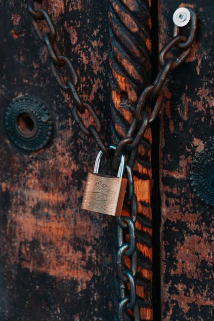 Close-up of a wooden door with a dark, weathered surface that has visible peeling paint and scratches. A metal chain with large, sturdy links is wrapped around the door handle area, secured with a brass padlock. The chain and padlock are essential for securing the door, possibly during a home relocation or storage process. The door features a glass peephole and metal fasteners, with a glimpse of an interior space or hallway behind it. In the context of house removals or packing and moving services, such security measures are often used to ensure safety during the packing and loading process before transportation by a van, which is typical of [COMPANY_NAME]'s service offerings. The environment appears inside an enclosed area, highlighting the importance of secure access during the moving logistics.