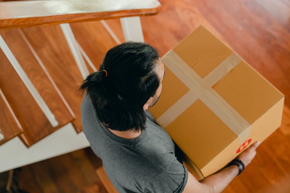 A person with dark hair tied back in a ponytail, wearing a grey T-shirt, is lifting a medium-sized cardboard box wrapped with beige packing tape, inside a home. The box features a red recycling symbol on one side. They appear to be navigating the box out of a room with wooden flooring and a white staircase railing visible in the background. The scene likely depicts a home relocation process, with the individual preparing to load or unload boxes as part of house removals and furniture transport. The lighting is natural, and the focus is on the careful handling of the packed box, emphasizing packing and moving activities coordinated by Man with Van Enfield Lock, operating within the removals service category.