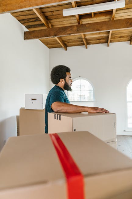A man with dark curly hair and a beard, wearing a blue t-shirt, stands inside a bright, spacious room with white walls, preparing for a house relocation. He is resting his hands on top of a large cardboard moving box with red packing tape, which appears to be part of a larger stack of packed boxes arranged on the floor. Behind him, there are two more cardboard boxes, one of which is smaller and placed atop a larger one. The room features a wooden ceiling with visible beams and a sloped roof, along with two arched windows allowing natural light to fill the space. The windows reveal an urban outdoor environment. The scene depicts the loading process of furniture and packing materials, with the focus on the careful handling and organization necessary for moving services provided by Man with Van Enfield Lock, as part of their house removals and furniture transport in the Enfield Lock area.