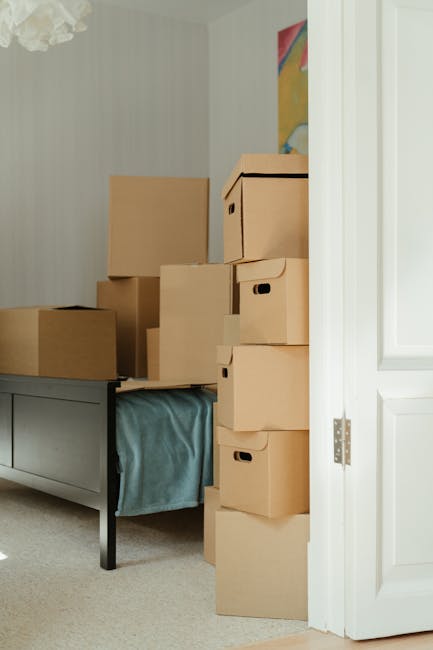 A person with dark hair tied back in a ponytail, wearing a grey T-shirt, is lifting a medium-sized cardboard box wrapped with beige packing tape, inside a home. The box features a red recycling symbol on one side. They appear to be navigating the box out of a room with wooden flooring and a white staircase railing visible in the background. The scene likely depicts a home relocation process, with the individual preparing to load or unload boxes as part of house removals and furniture transport. The lighting is natural, and the focus is on the careful handling of the packed box, emphasizing packing and moving activities coordinated by Man with Van Enfield Lock, operating within the removals service category.