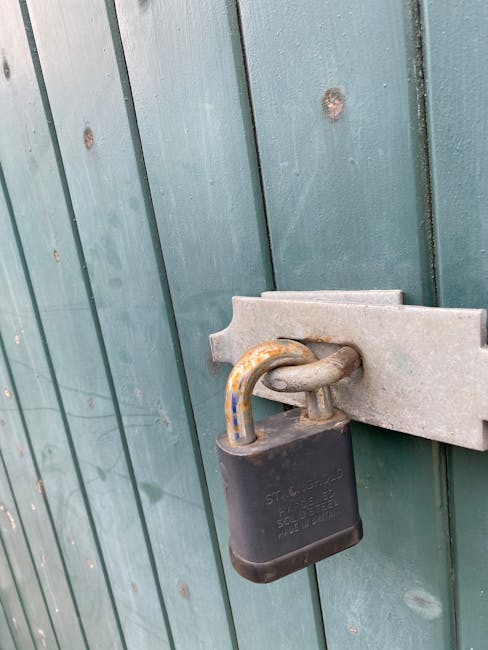 Close-up view of a black padlock securing a metal latch attached to a green wooden fence. The fence panels are vertically aligned with visible nails and a weathered finish. The padlock's shackle shows signs of rust and corrosion, and the lock is positioned at the center of the image, indicating it is used to secure an entrance or boundary. This image is related to house removals and home relocation services, illustrating the importance of securing properties during moving or transportation activities, as provided by Man with Van Enfield Lock near Enfield Lock station, ENFIELD LOCK.