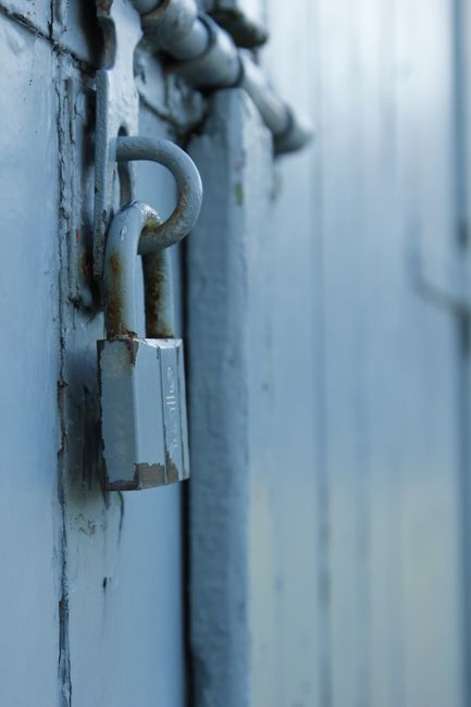 Close-up image of a metal padlock securing a blue-painted wooden door, with a rusted, weathered padlock and sturdy metal hasp. The door features visible vertical wooden planks with chipped and peeling paint, indicating age and exposure to the elements. The padlock and hasp are attached to the door, suggesting it is locked and secured. The scene is illuminated with natural light, highlighting the texture of the weathered paint, rust on the lock, and the rough surface of the wood. This image relates to house removals and secure storage, emphasizing the importance of secure closures during packing, moving, and transportation processes, as managed by companies like Man with Van Enfield Lock.