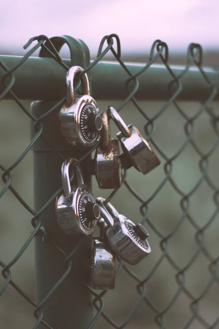 Close-up of a black metal chain-link fence with three silver combination padlocks attached, each securing the gate. The padlocks are different sizes, with one larger and two smaller, and are positioned near the upper left section of the fence. The background is blurred, showing an outdoor environment with a pastel sky and distant greenery, indicating an exterior setting. The image emphasizes security and locking mechanisms, which can relate to safeguarding belongings during house removals or the transportation phase of home relocation services provided by Man with Van Enfield Lock.