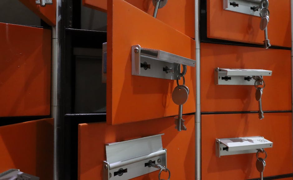 Close-up view of orange communal mailboxes with silver keyholes and horizontal locking bars, each mailbox secured with a key hanging from a metal latch. The mailboxes are mounted on a wall in an indoor setting, with some open showing dark interiors. The lighting appears to be artificial, creating reflections on the metal surfaces. This image captures the area typically used in residential or commercial buildings for mail collection, with a focus on security features like keys and locks. It illustrates the importance of proper access control during home relocation or moving processes, as part of packing and planning for furniture transport or access to building entry points, which Man with Van Enfield Lock facilitates through professional removals services.